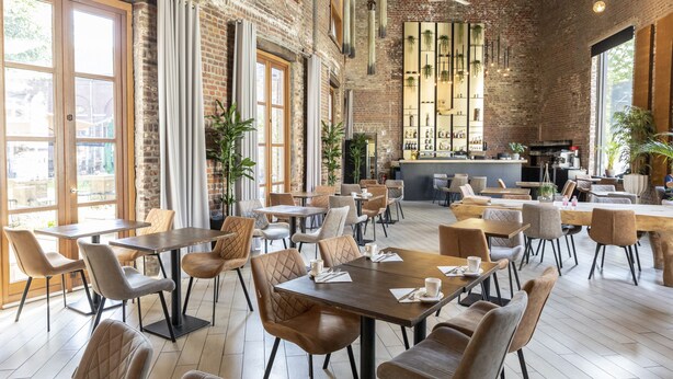 A spacious café with brick walls, large windows, and a variety of seating arrangements including tables and chairs. - Eburon Hotel - Different Hotels (Tongeren)