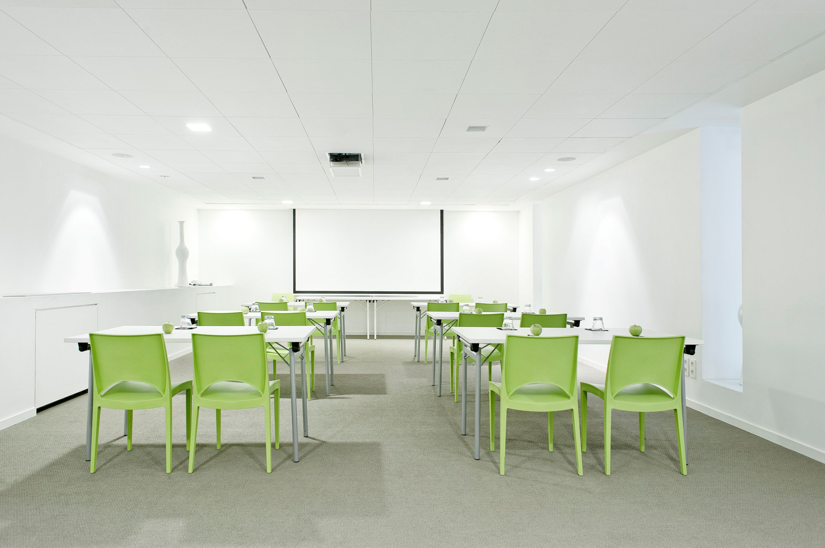 A conference room with white walls, a projector screen, and green chairs arranged around a central table.