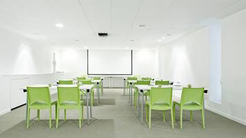 A conference room with white walls, a projector screen, and green chairs arranged around a central table.