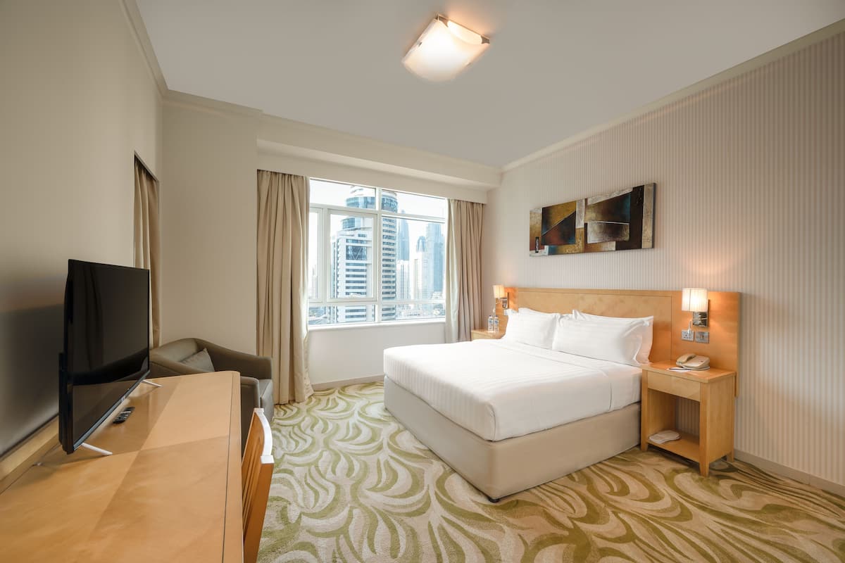 Habitación de hotel con una cama grande, un escritorio con televisor, una mesita de noche y una ventana con cortinas.