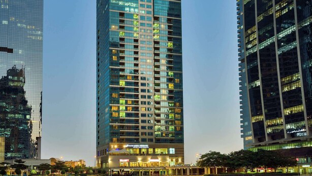 A modern high-rise building with a glass facade, situated next to a body of water reflecting its lights. - Oaks Liwa Heights Suites (Dubai)