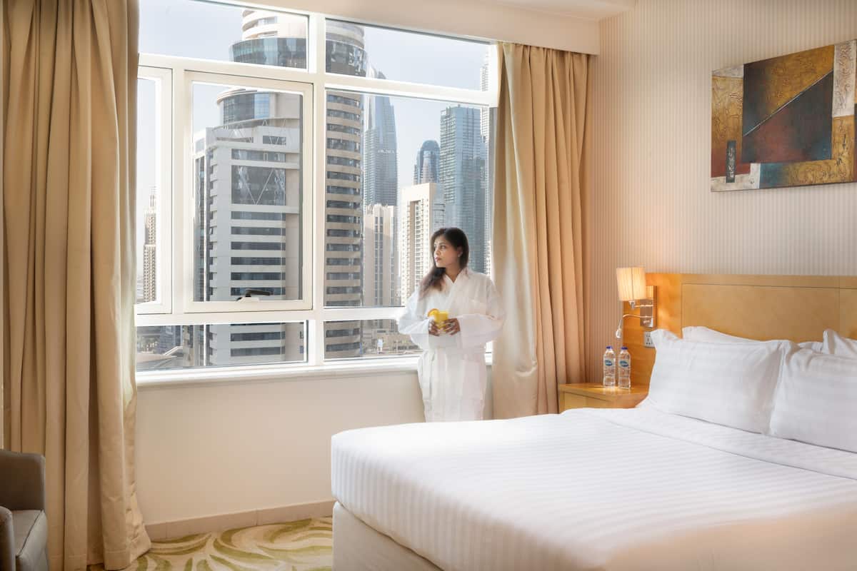 Habitación de hotel con una cama grande, una ventana con cortinas, una mesita de noche con lámpara y un cuadro en la pared.