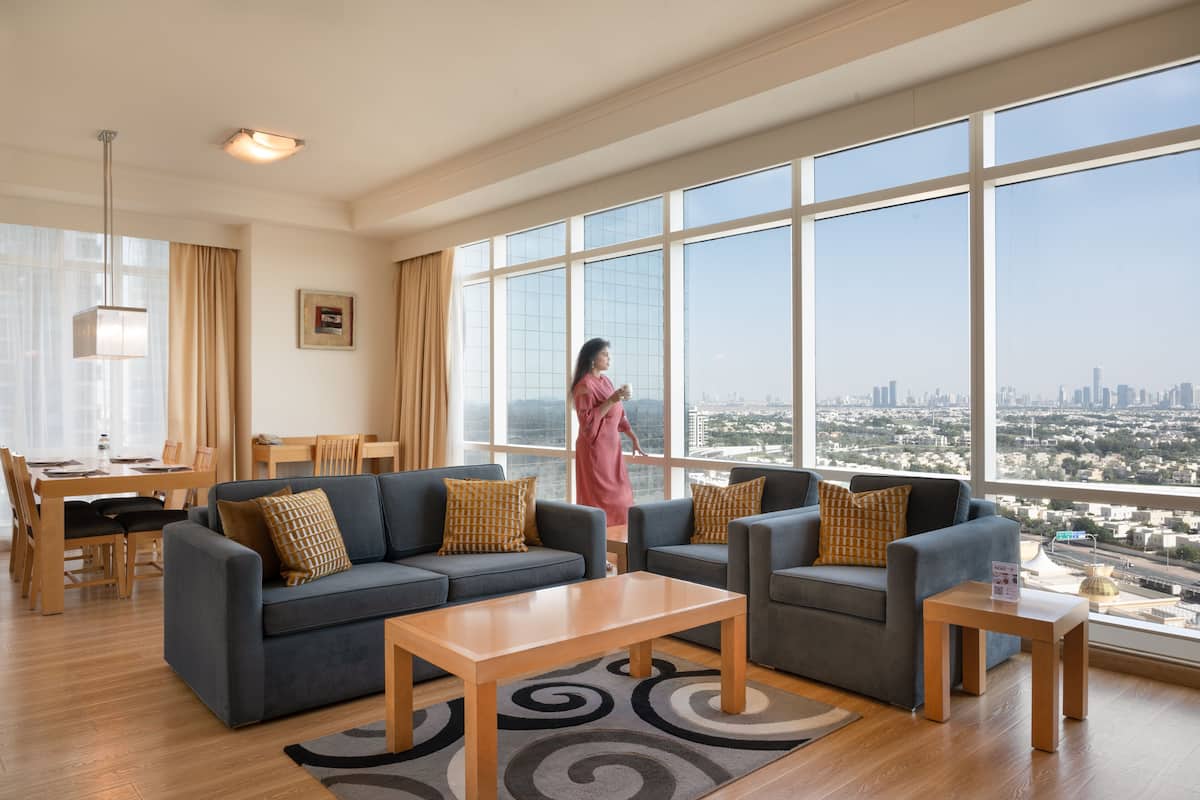 Amplia sala de estar con un gran ventanal que ofrece vistas a la ciudad, dos sofás con cojines estampados, una mesa de centro de madera y una zona de comedor con mesa y sillas.