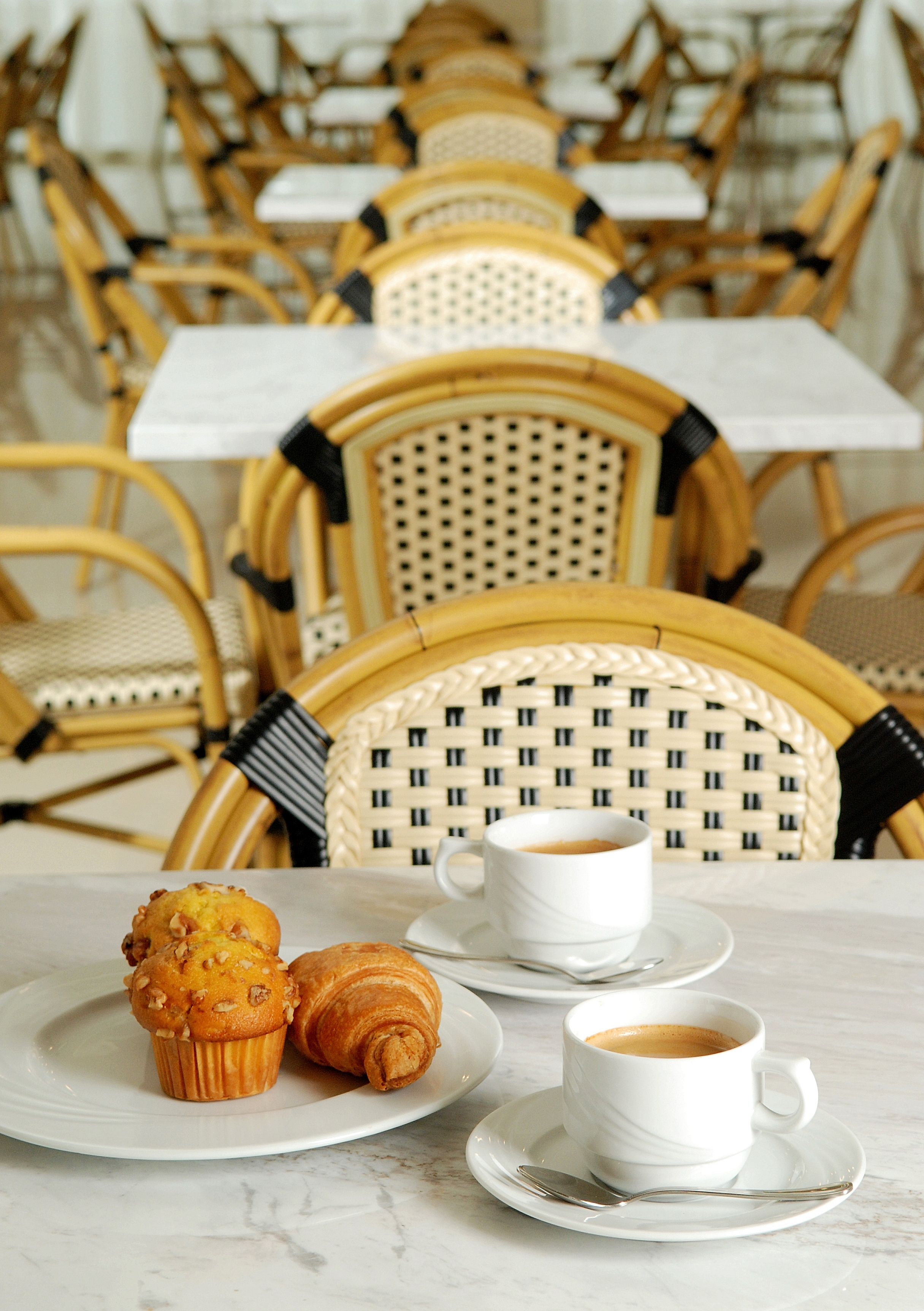 Un coin repas avec des chaises et des tables en osier, une assiette contenant un muffin et un croissant, et une tasse de café.