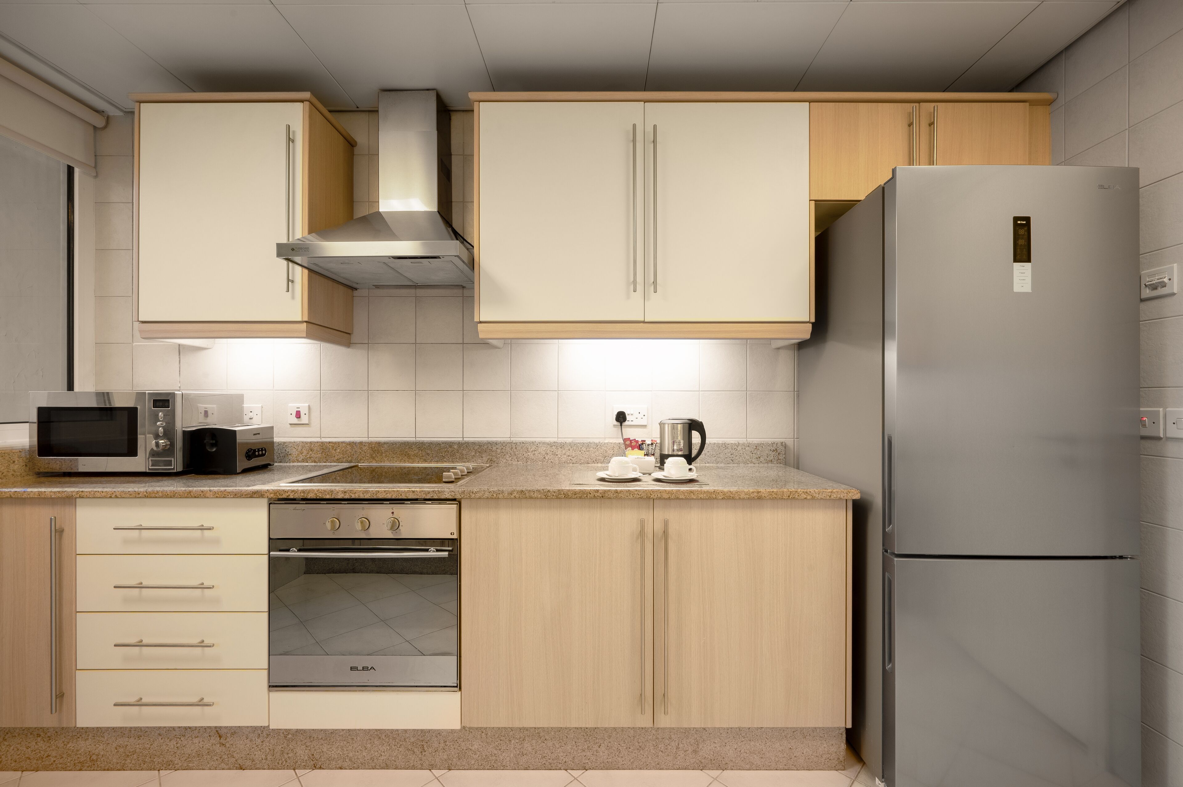 A modern kitchen with light wood cabinets, a stainless steel refrigerator, a microwave, an oven, and a coffee maker.