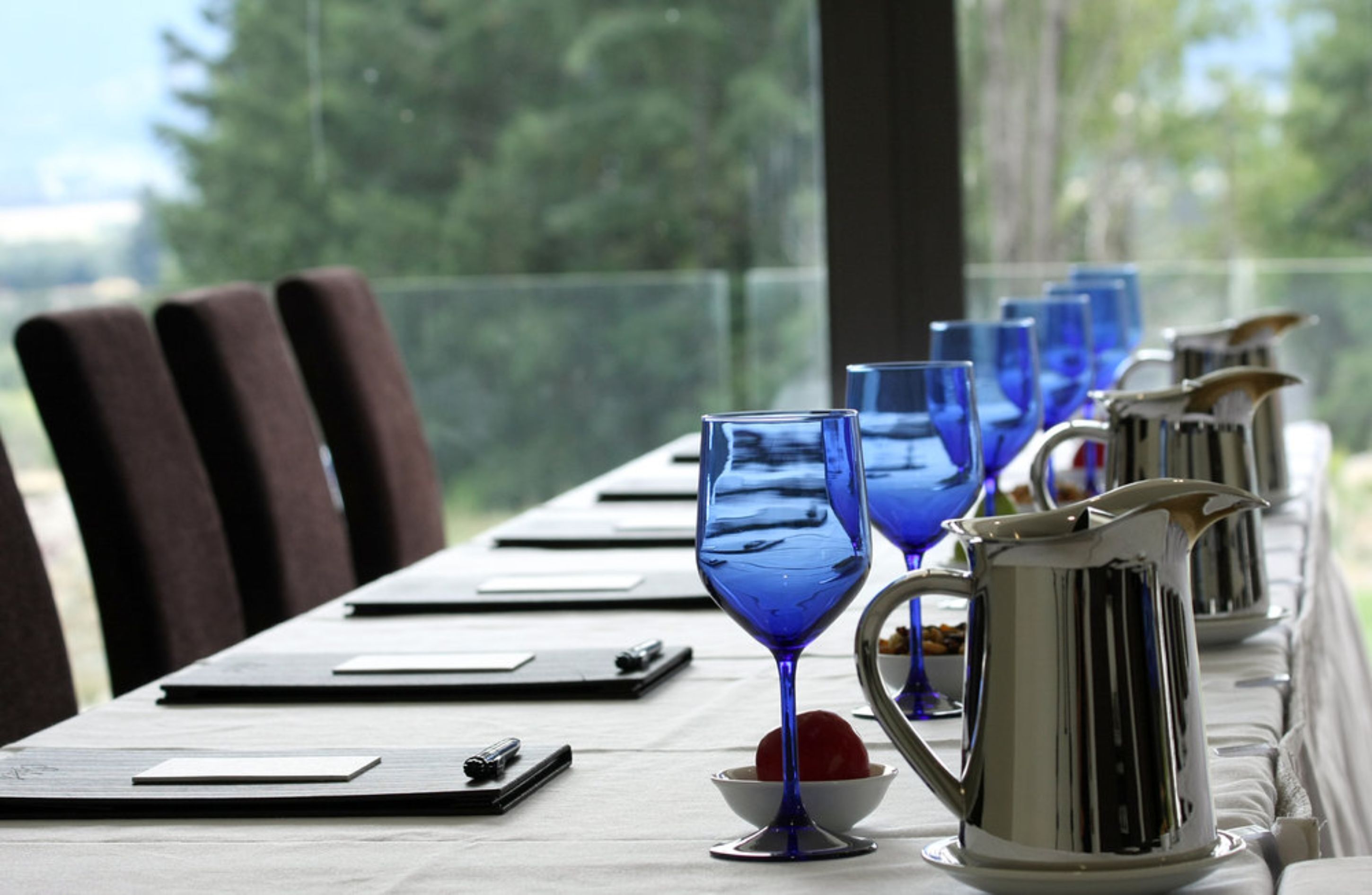Une table à manger avec des verres à vin bleus et un pichet en argent, le tout mis en valeur par un décor composé de chaises et offrant une vue sur des arbres.