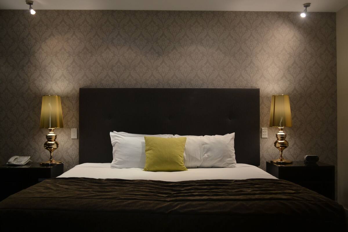 A neatly made bed with a dark headboard, two bedside lamps, and a patterned wallpaper.