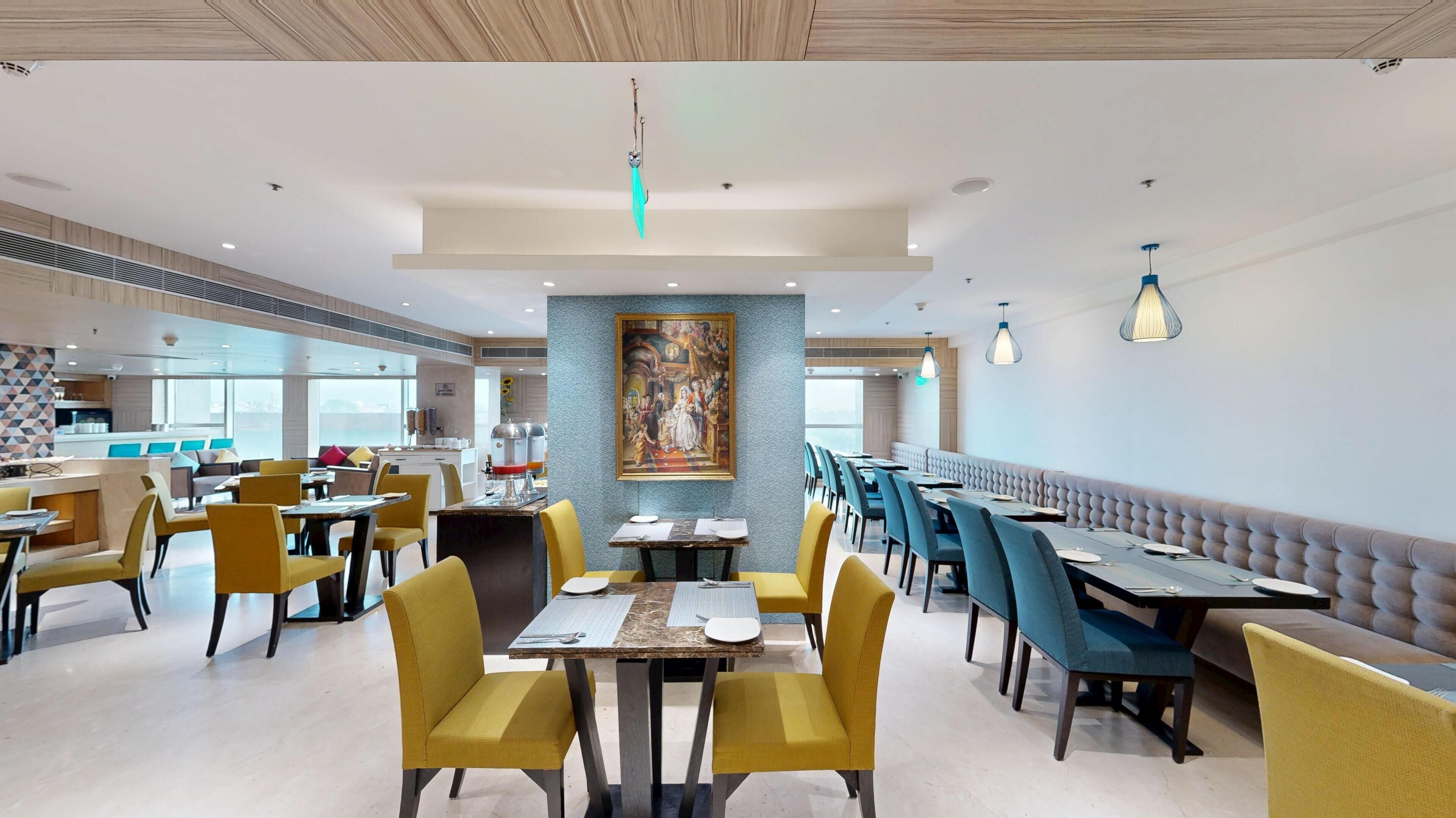 Un ristorante moderno con sedute gialle e blu, un murale centrale e una cucina visibile sullo sfondo.