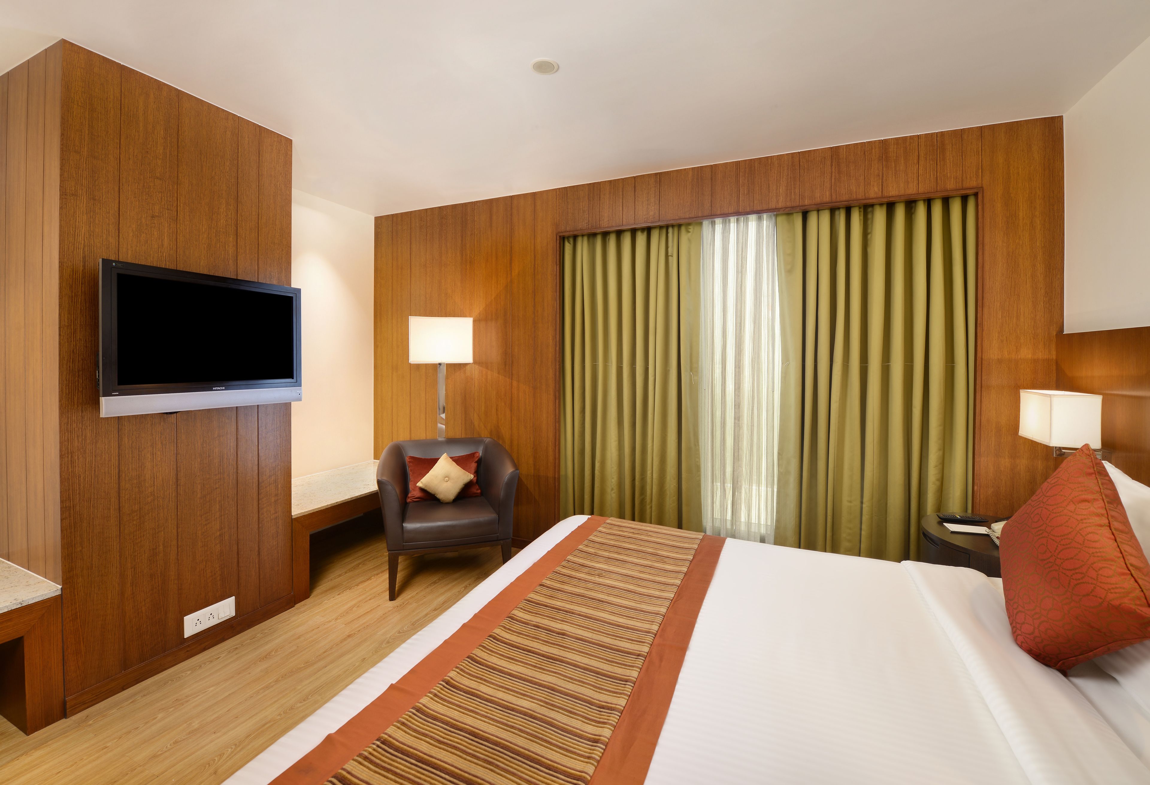 A hotel room with a large bed, a flat-screen TV mounted on the wall, a desk with a lamp, and a chair with a pillow.