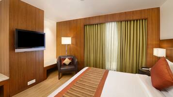 A hotel room with a large bed, a flat-screen TV mounted on the wall, a desk with a lamp, and a chair with a pillow.