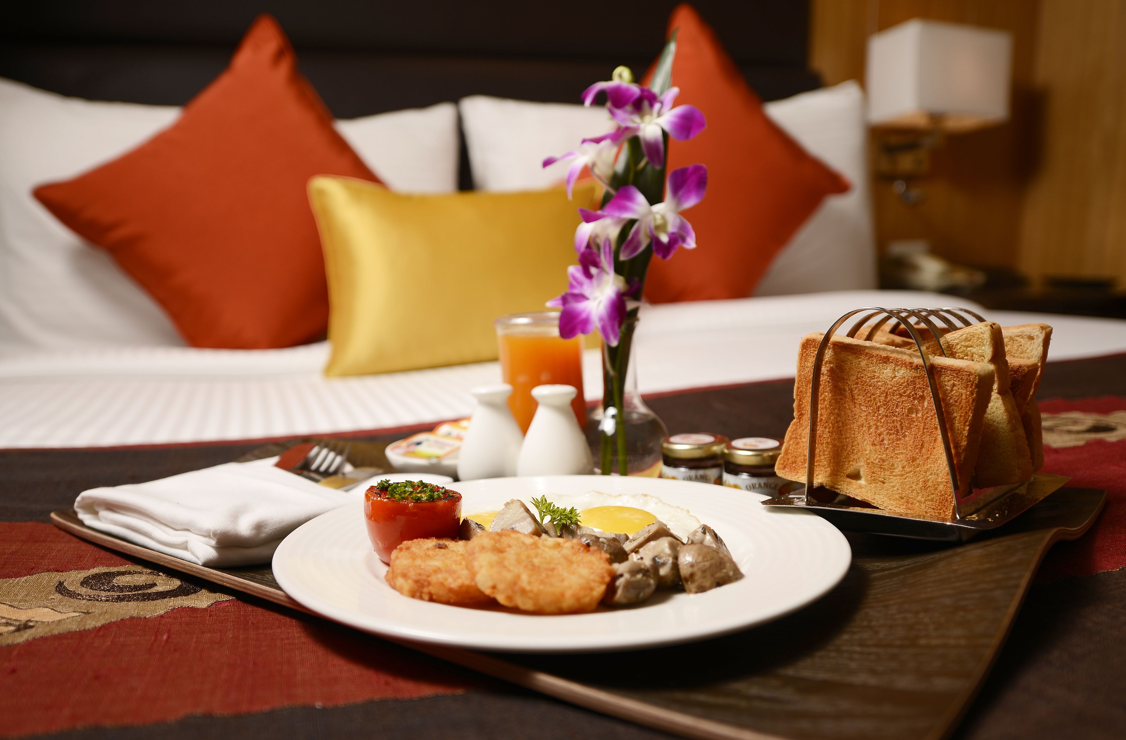 A breakfast setting with a plate of food, a vase with flowers, and a bed with pillows in the background.