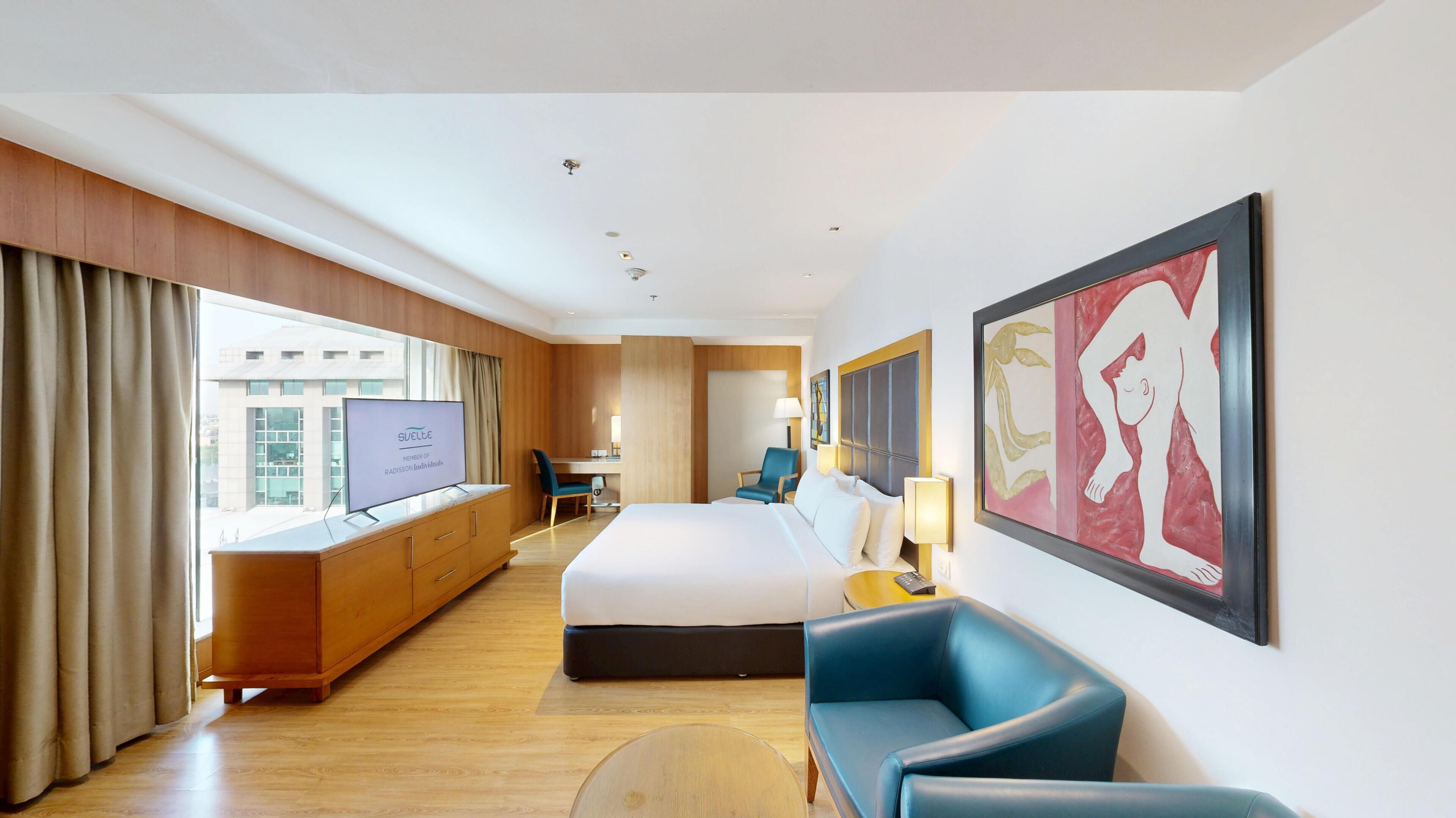 A hotel room with a bed, a desk, a chair, a TV, and artwork on the wall.