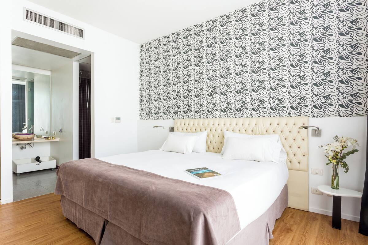 A modern hotel room with a large bed, a tufted headboard, a bedside table with a vase of flowers, and a patterned wall.