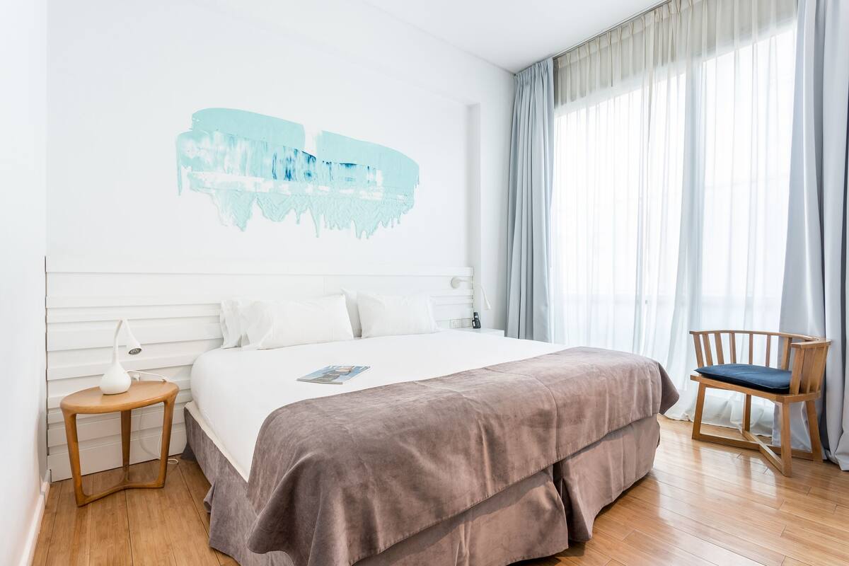 A modern hotel room with a large bed, a bedside table, a chair, and a window with curtains.
