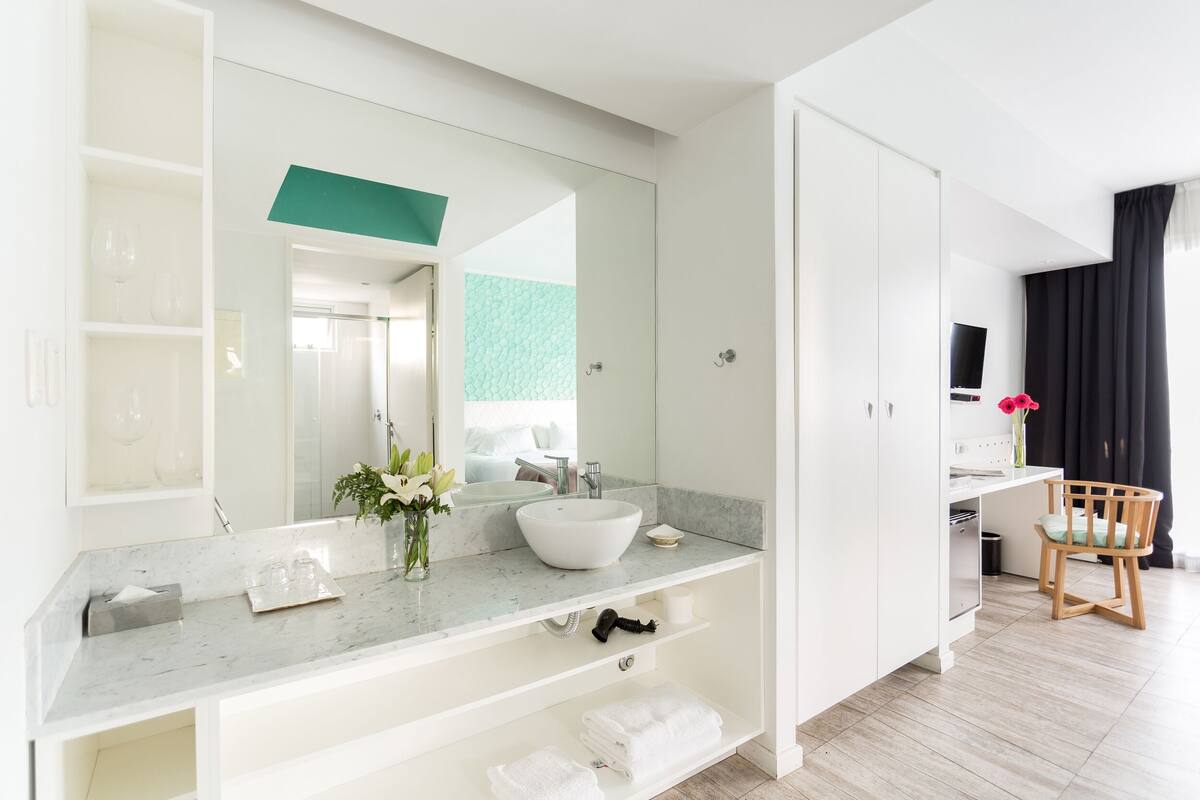 A modern bathroom with a large mirror, white vanity, and a sink. There is a wooden chair and a small table with a vase of flowers.