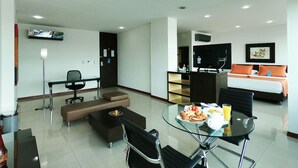 Quarto de hotel moderno com mesa de jantar de vidro, sofá, escrivaninha e cama com almofadas laranja.