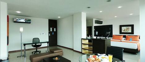 A modern hotel room with a glass dining table, a sofa, a desk, and a bed with orange pillows.