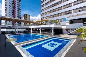 Una piscina en la azotea con una fuente de agua clara y azul, rodeada de edificios modernos y zonas de estar al aire libre.