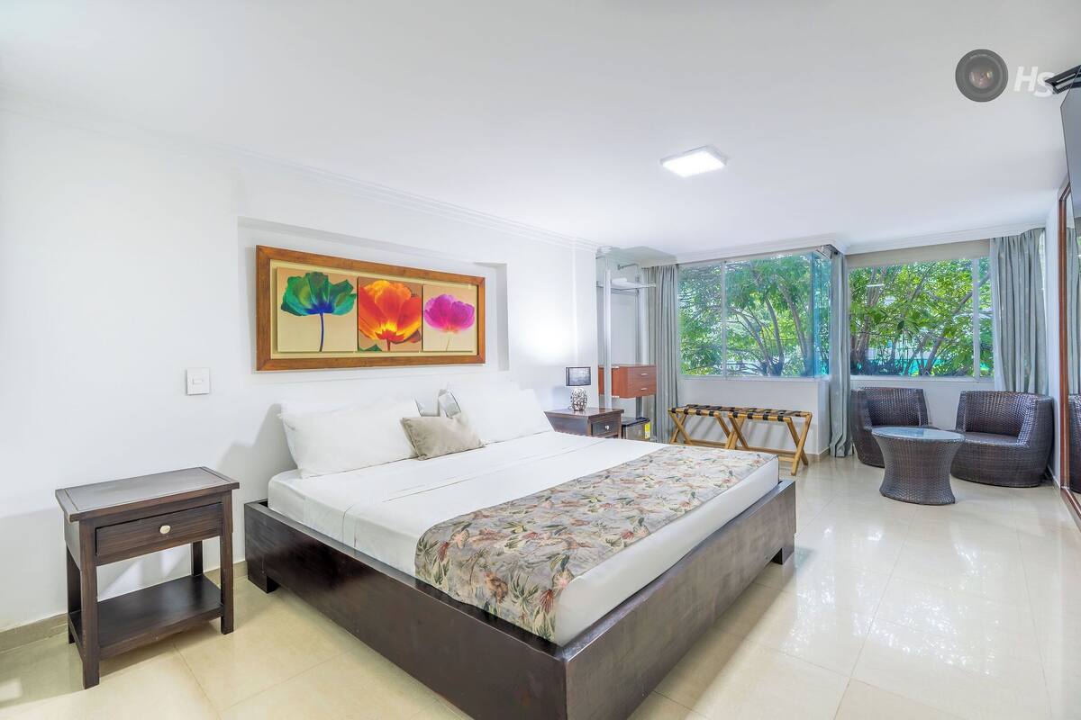 A modern hotel room with a large bed, a small side table, a wicker sofa, and a large window offering a view of greenery.