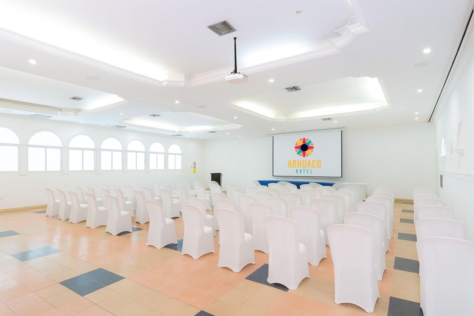 Une salle de conférence spacieuse, avec des rangées de chaises blanches, un pupitre et un écran de projection affichant le logo de l’hôtel Arhuaco.