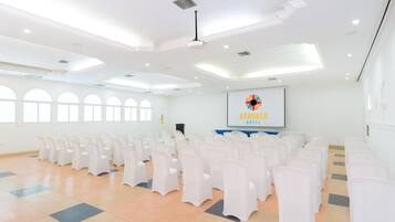 Une salle de conférence spacieuse, avec des rangées de chaises blanches, un pupitre et un écran de projection affichant le logo de l’hôtel Arhuaco.
