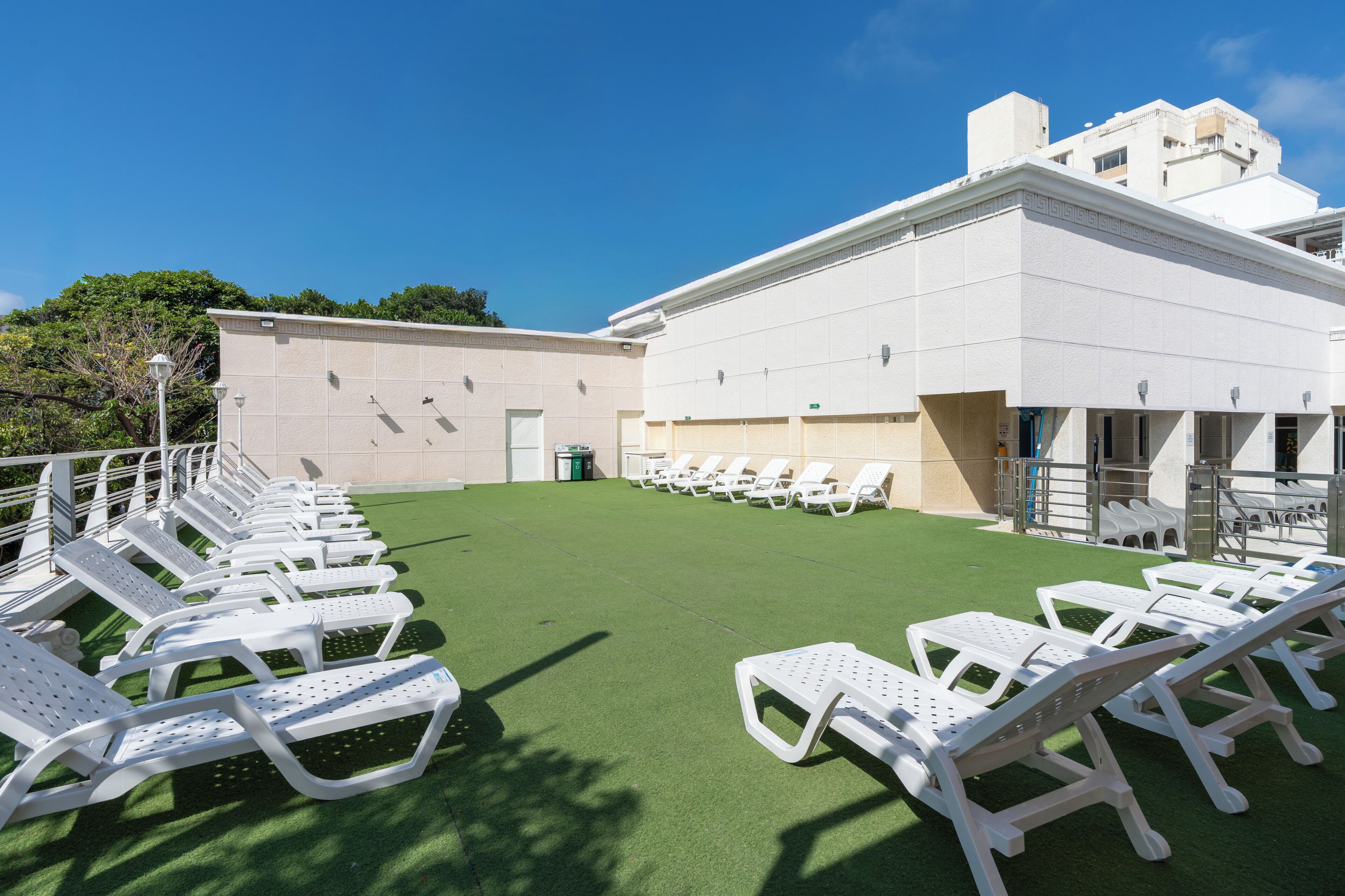 Una terraza en la azotea con sillones blancos de sala dispuestos en filas, césped artificial verde y un edificio blanco con techo plano y fachada color crema.