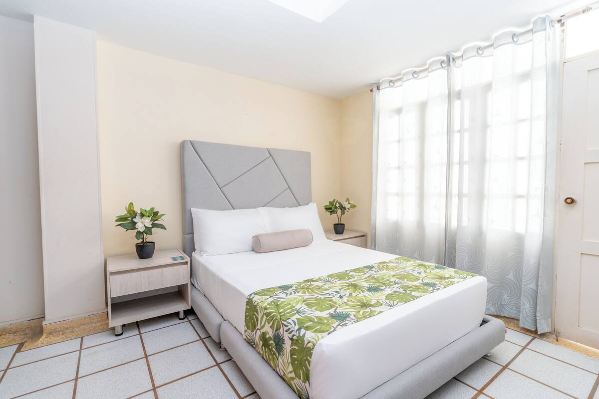 A bedroom with a bed, a nightstand, a potted plant, and a window with curtains.