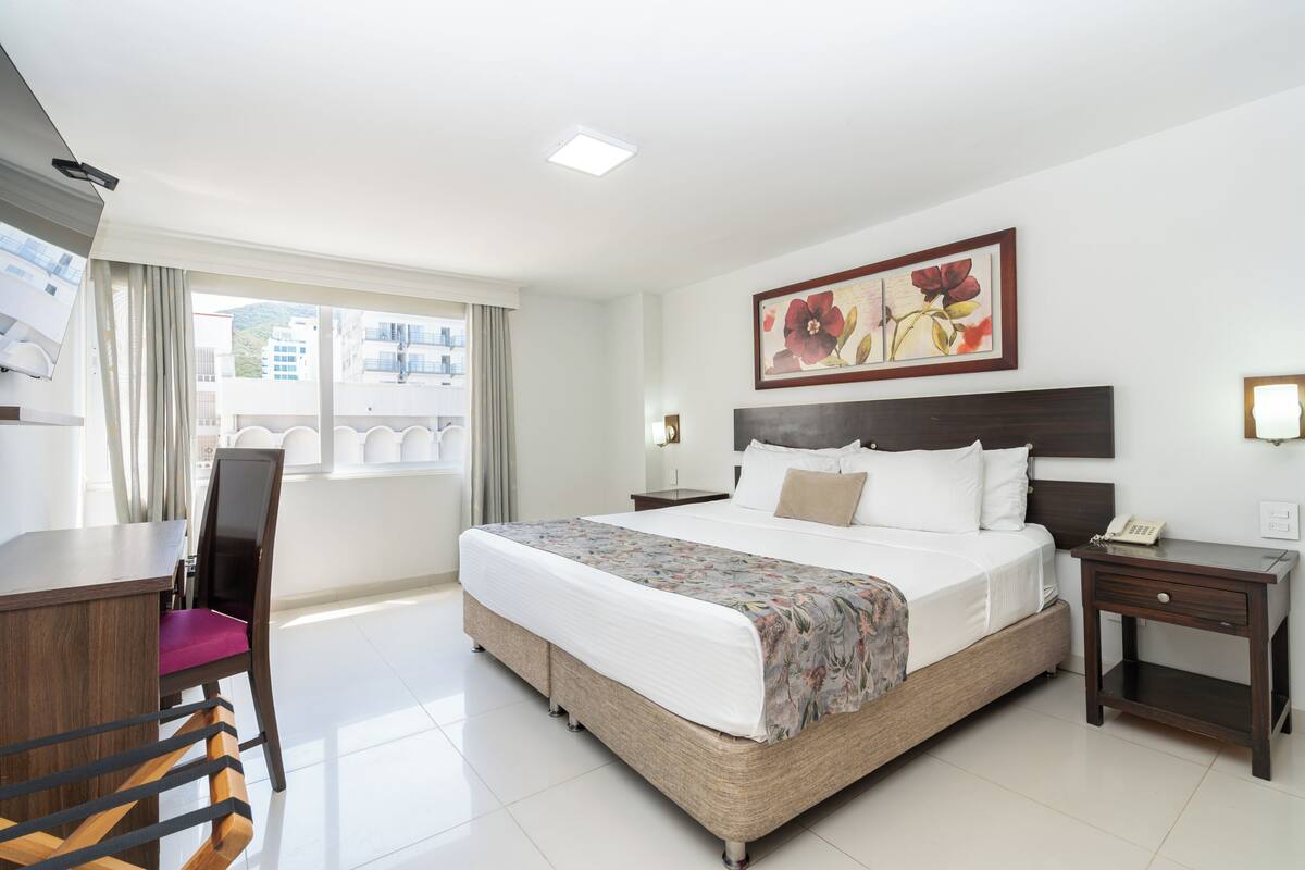 A hotel room with a bed, a desk, a chair, a bedside table, and a window with curtains.