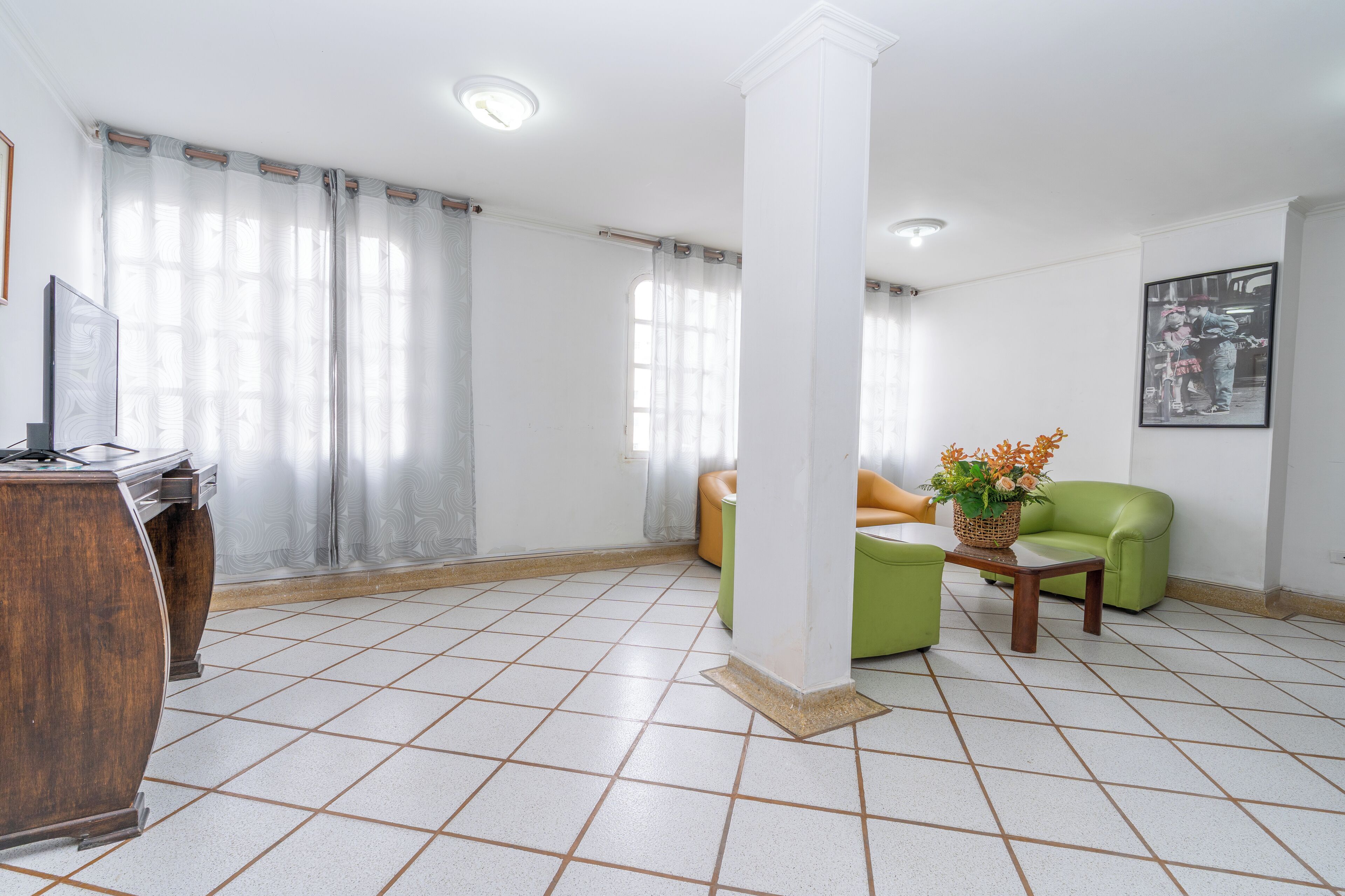 A spacious living area with a tiled floor, a wooden desk, a green sofa, and a small table with a flower vase.