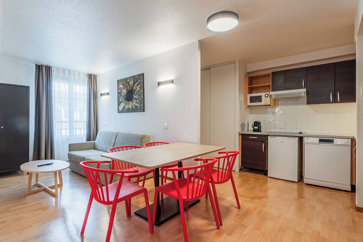 A modern living room with a dining table, red chairs, a grey sofa, and a kitchen area with cabinets and appliances.