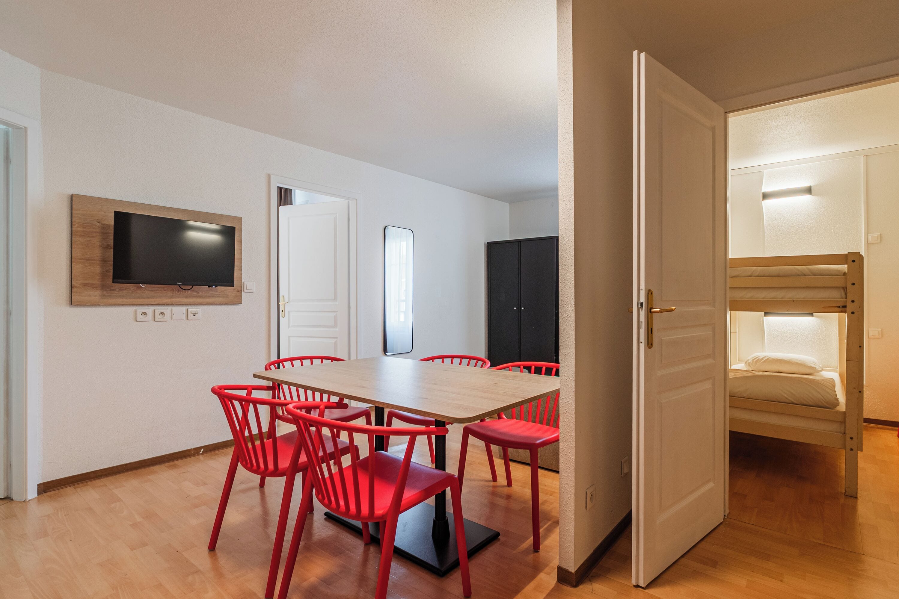 A room with a wooden dining table, red chairs, a flat-screen TV mounted on the wall, a door leading to a bedroom with a bunk bed, and a black cabinet.