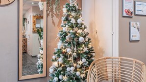 Een kerstboom met witte versieringen en lichtjes, een rieten stoel, een spiegel, en een muur met informatieposters.