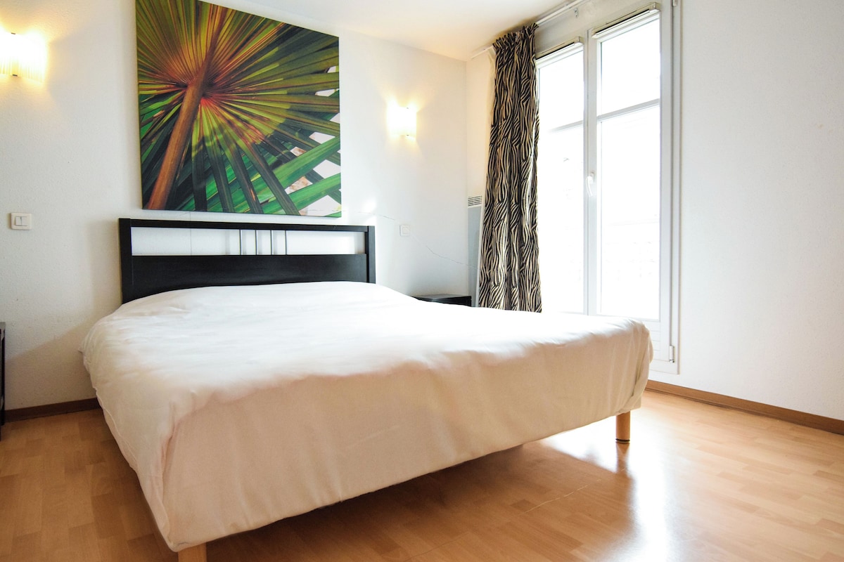 Un dormitorio con cama, una obra de arte en la pared, ventana con cortinas y una lámpara.