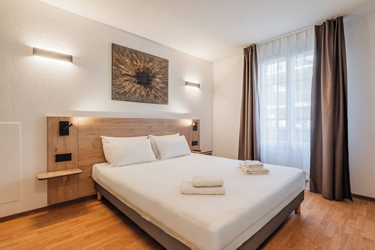 A modern hotel room with a large bed, wooden headboard, bedside tables, and a wall-mounted artwork.