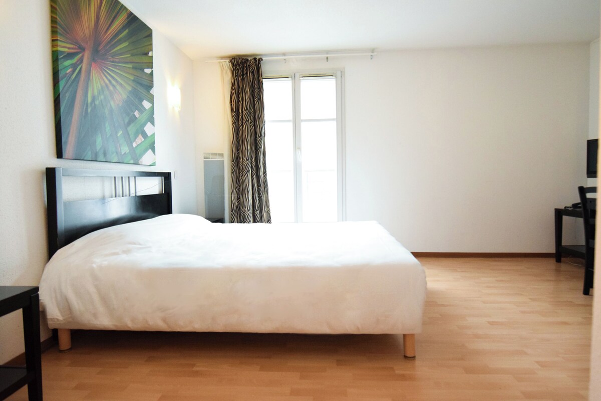 Um quarto com uma cama grande, cabeceira, janela com cortinas e uma obra de arte pendurada na parede.