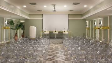 Un salón de conferencias con sillas transparentes dispuestas en filas, un atril con micrófonos y una pantalla grande en blanco al frente.