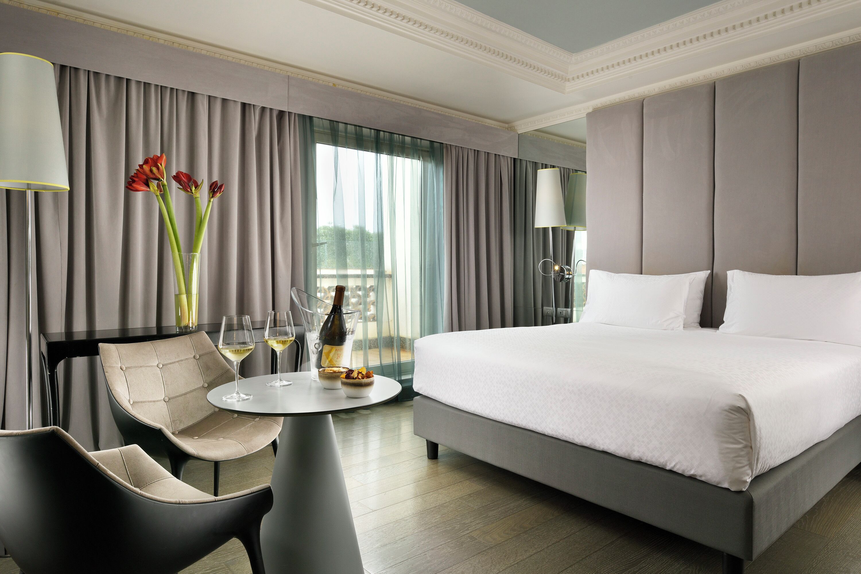 A modern hotel room with a large bed, a round table with two chairs, a vase with flowers, and a view of the outdoors through the window.
