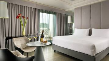 A modern hotel room with a large bed, a round table with two chairs, a vase with flowers, and a view of the outdoors through the window.