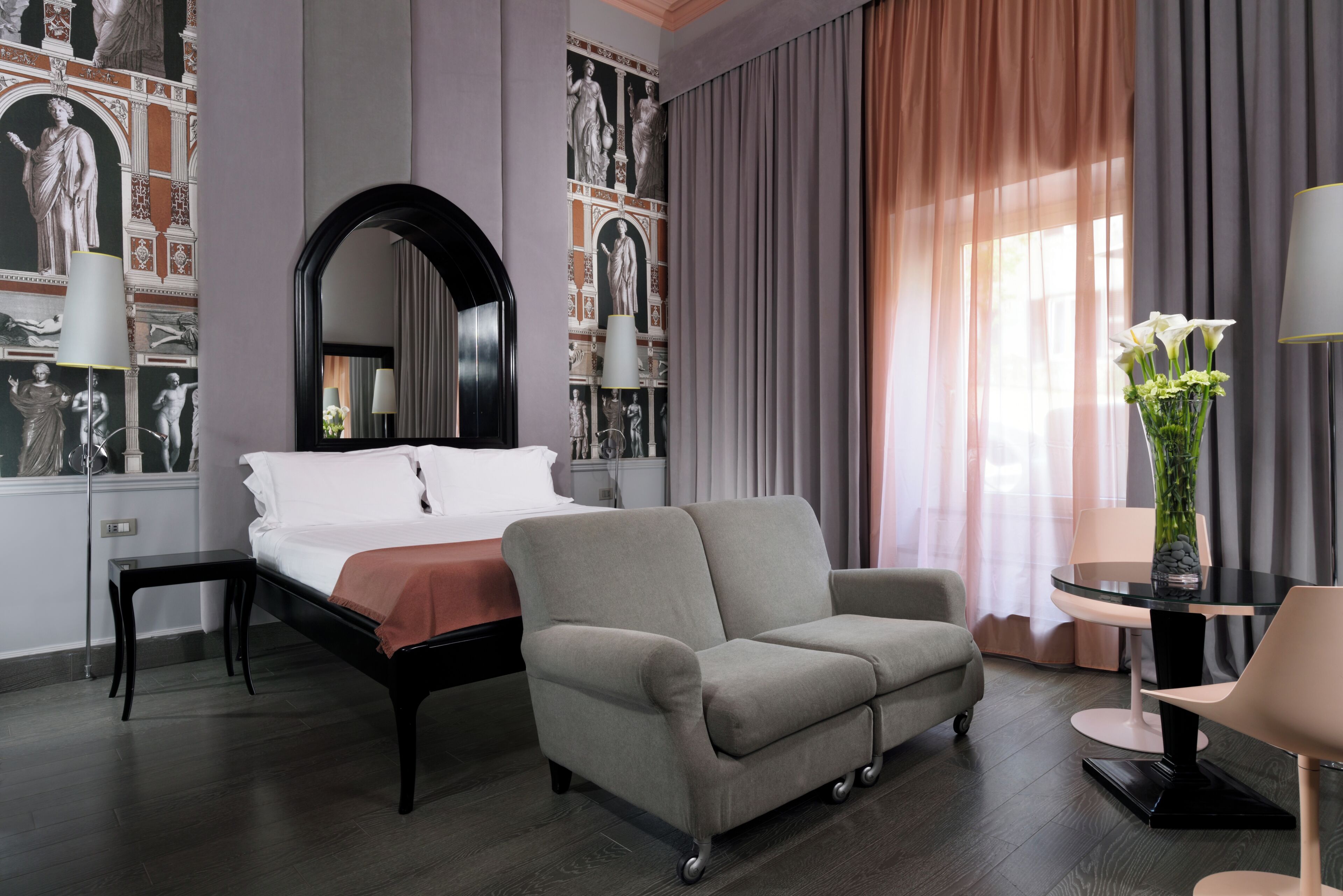 Een hotelkamer met een bed, een grijze fauteuil, een ronde tafel met een lamp en een vaas met bloemen.