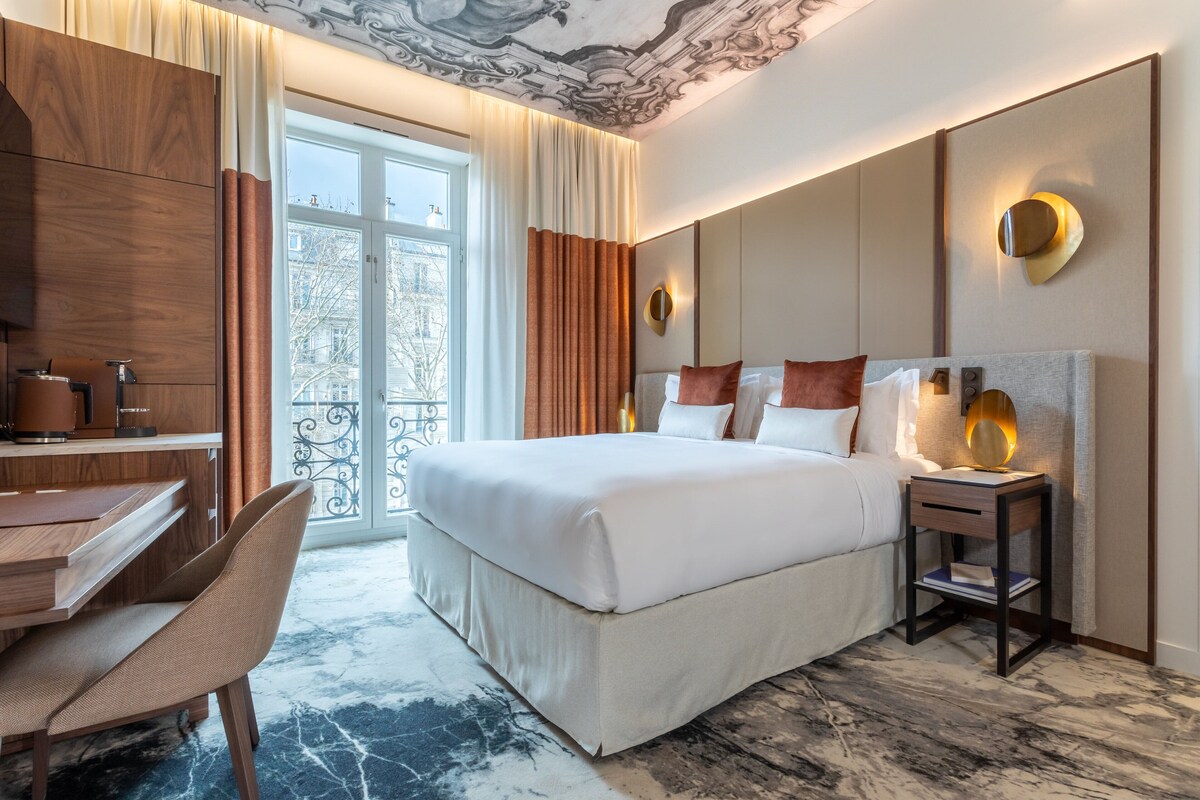 A hotel room with a bed, desk, chair, and a view of the outdoors.