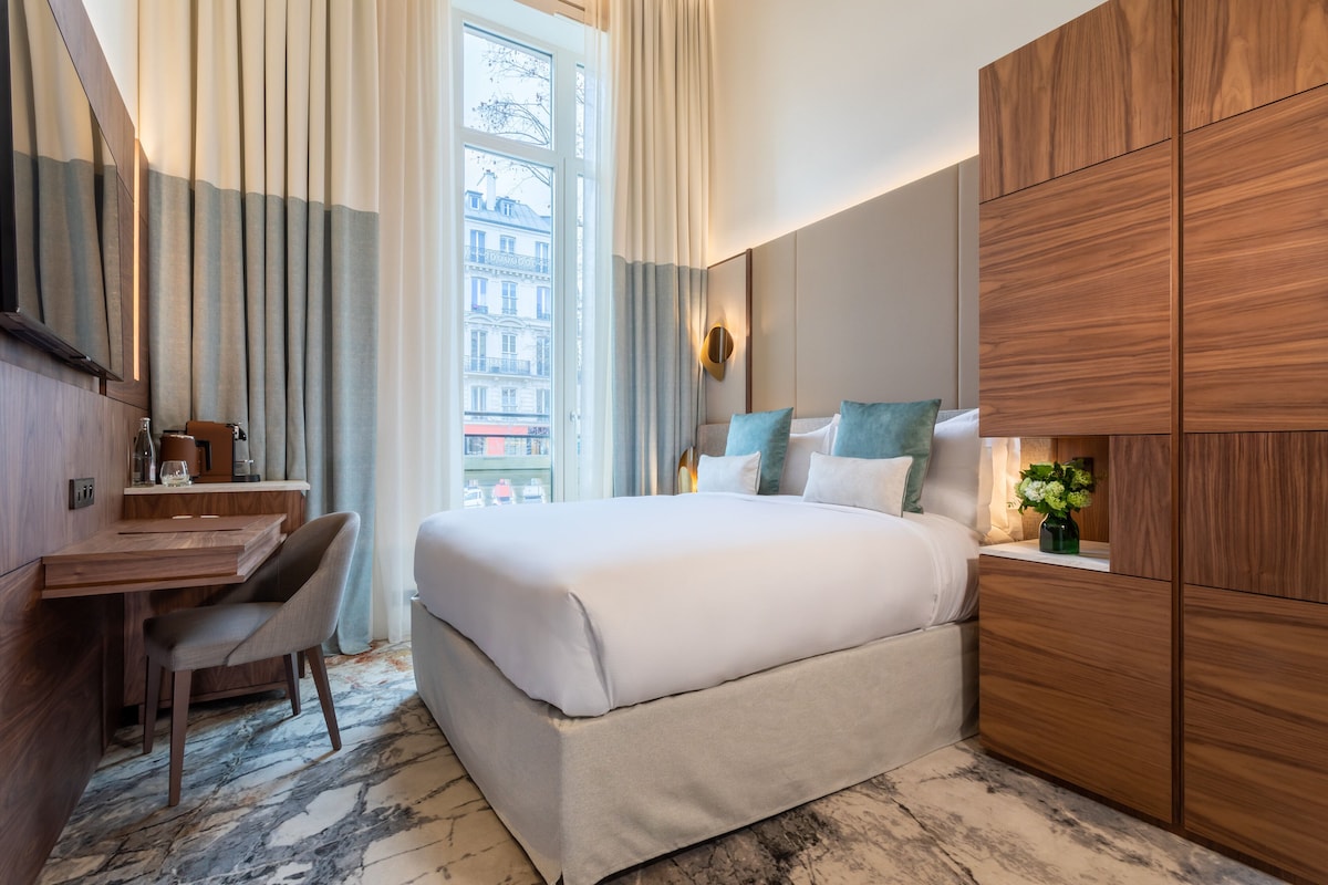 A modern hotel room with a large bed, wooden wardrobe, a desk with a chair, and a view of a building through the window.