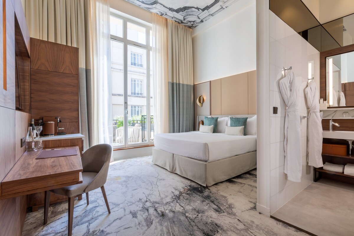 A hotel room with a bed, a desk, a chair, a bathroom with a mirror, and a view of the outside.
