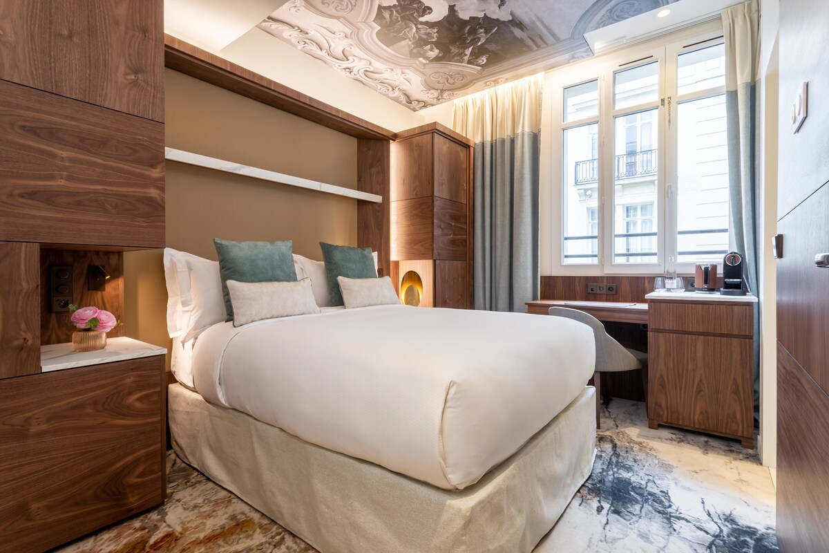 A modern hotel room with a large bed, wooden furniture, a desk, and a view of the city through large windows.