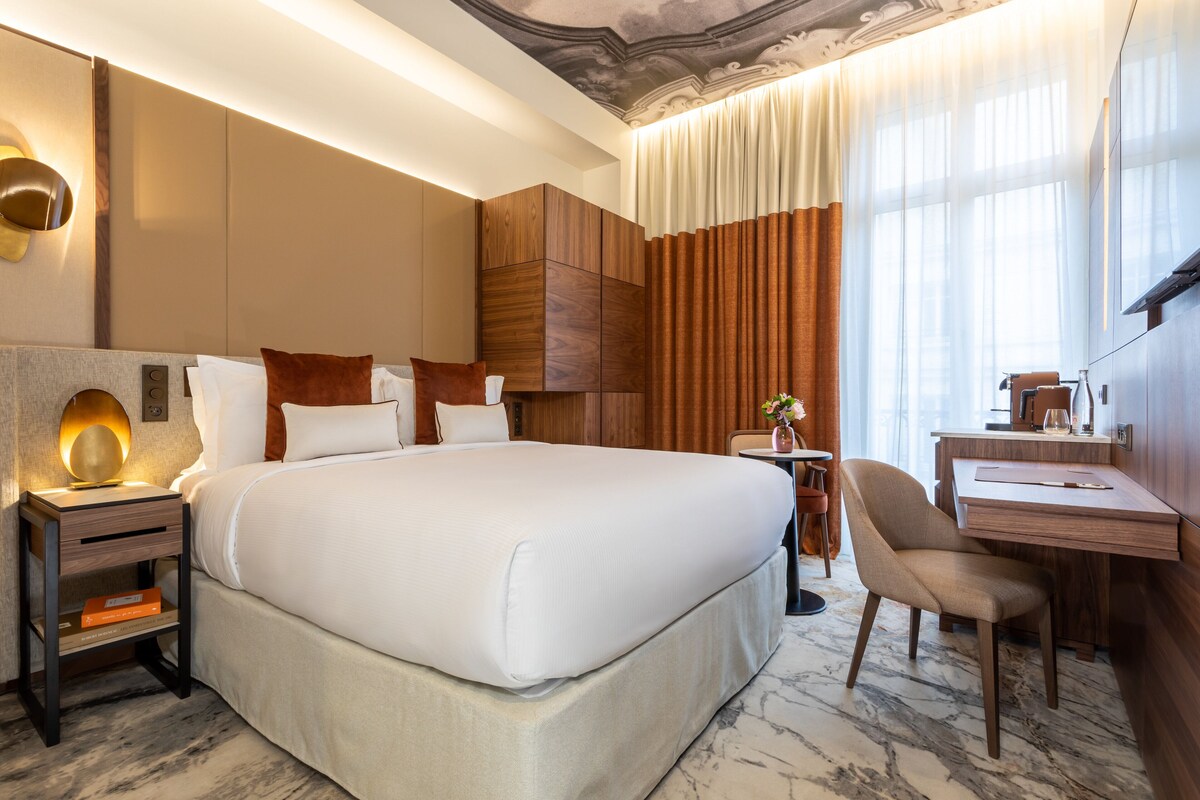 A hotel room with a large bed, a desk, and a chair. There is a nightstand with a lamp and a vase with flowers. The room has wooden paneling and a marble floor.