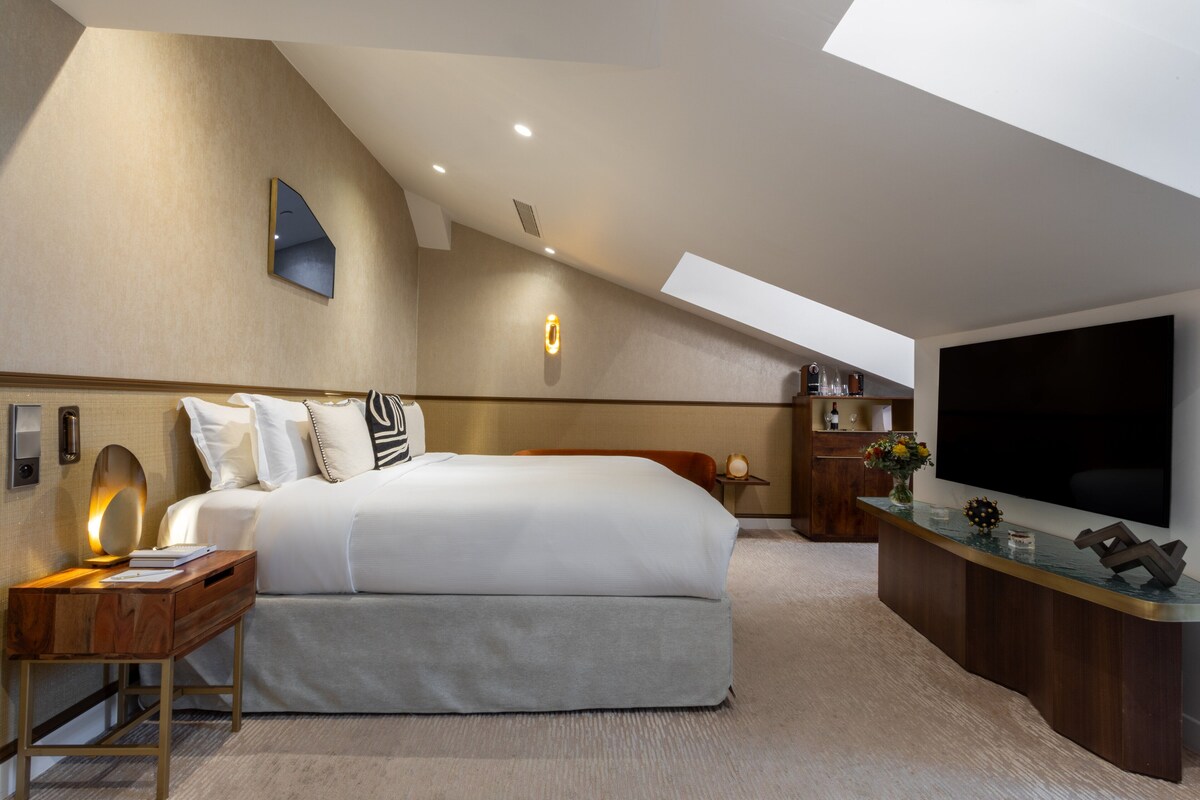 A modern hotel room with a large bed, a desk with a lamp, a TV, and a small side table.