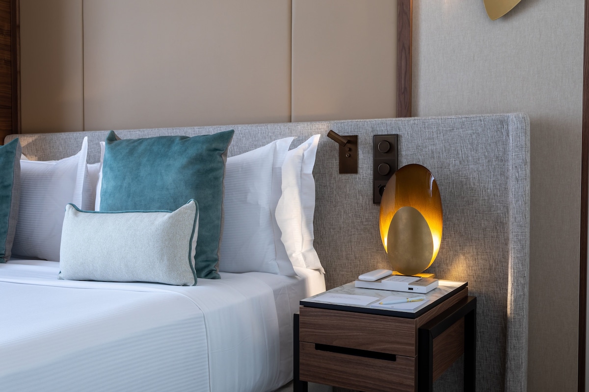 A neatly made bed with white and teal pillows, a bedside table with a lamp, and a wall-mounted light fixture.