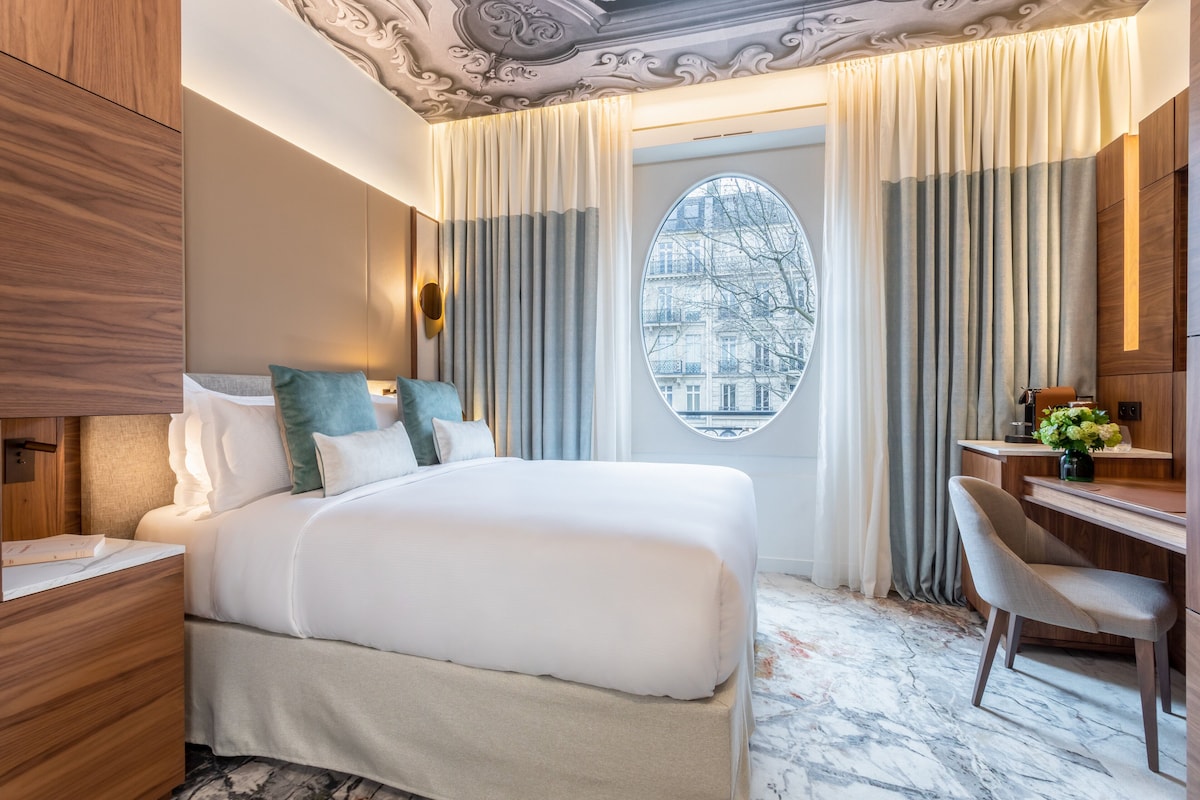 A hotel room with a large bed, a desk, and a view of a cityscape through a window.