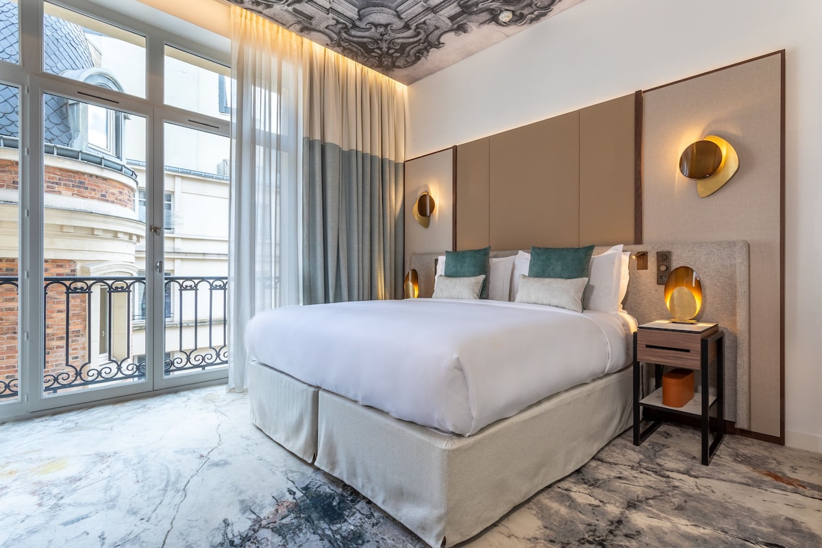 A hotel room with a large bed, a nightstand with a lamp, a view of a building through a window, and a balcony.