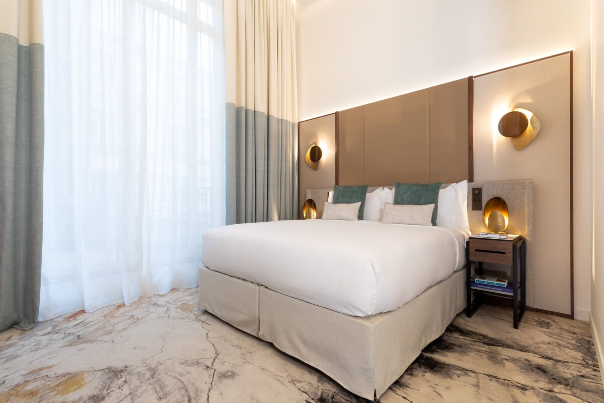 A hotel room with a large bed, a nightstand, and a wall-mounted light fixture.