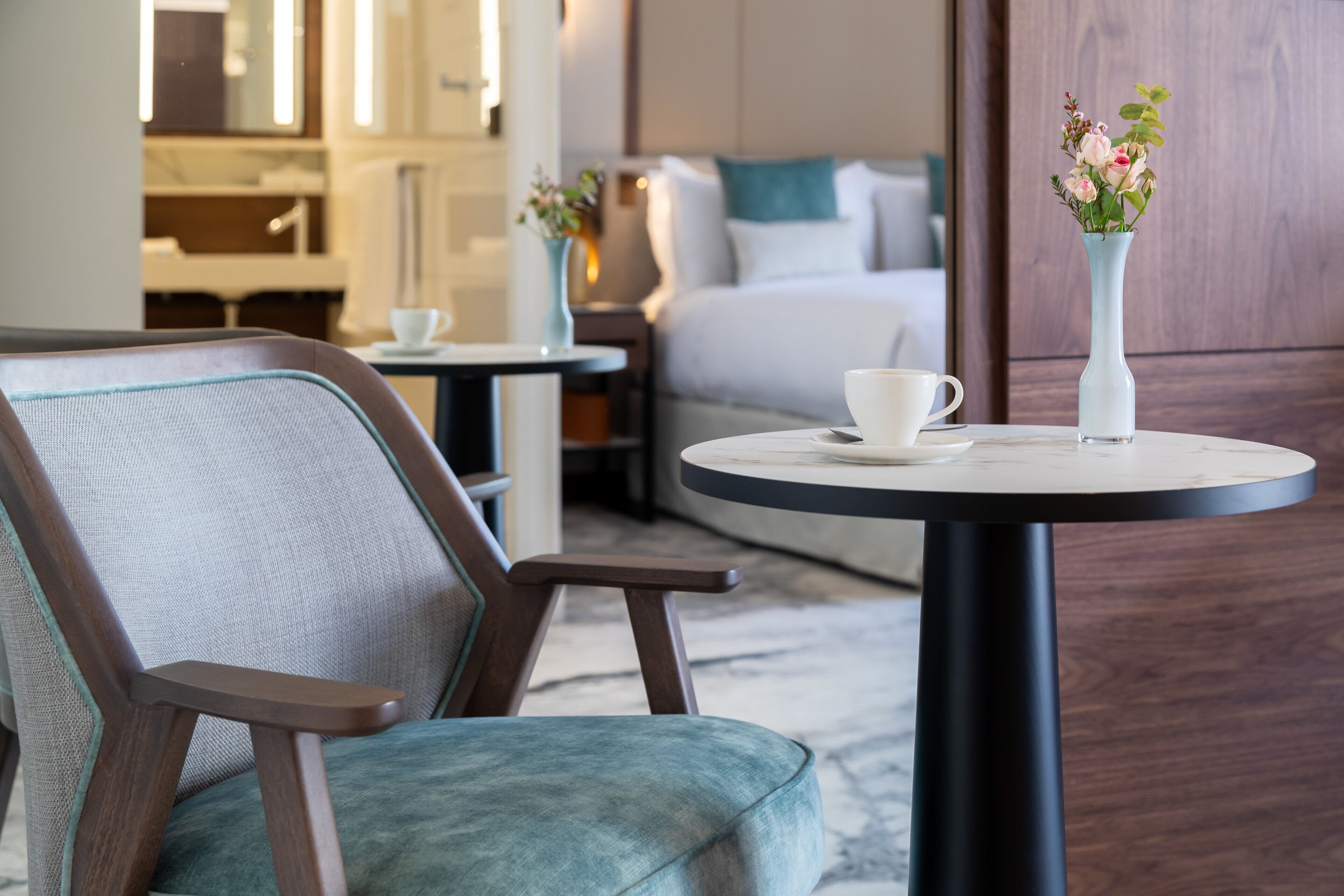 Una habitación de hotel moderna con una mesa redonda, un sillón cómodo, una cama con ropa de cama blanca y un jarrón con flores sobre la mesa.